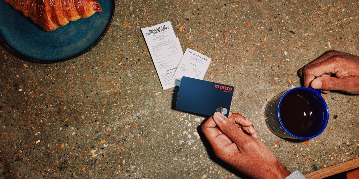 A business owner holds their Monzo Business card at a cafe counter with freshly baked croissant and coffee nearby.