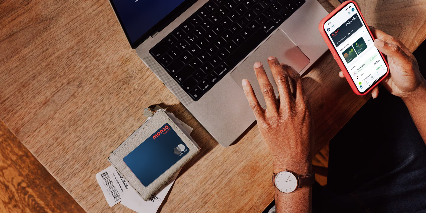 Overhead view of a business owner's workspace showing a laptop, phone displaying the Monzo app, and a Monzo business card in its holder on a wooden desk.