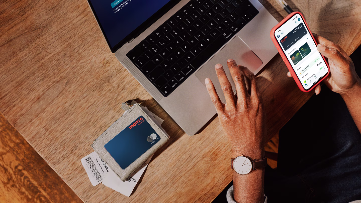 Overhead view of a business owner's workspace showing a laptop, phone displaying the Monzo app, and a Monzo business card in its holder on a wooden desk.