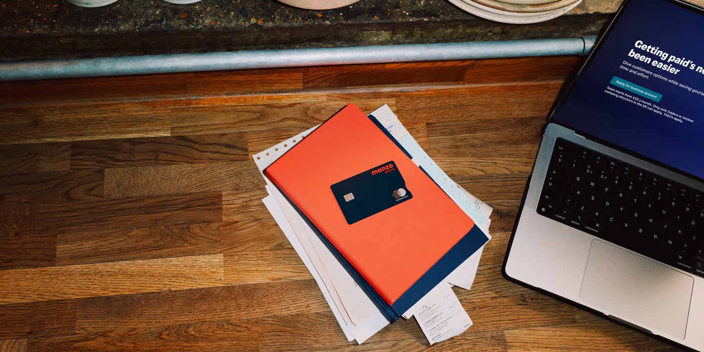 Overhead view of a restaurant workspace with a Monzo business card resting on paperwork beside a laptop, stacked plates visible on the shelf above.