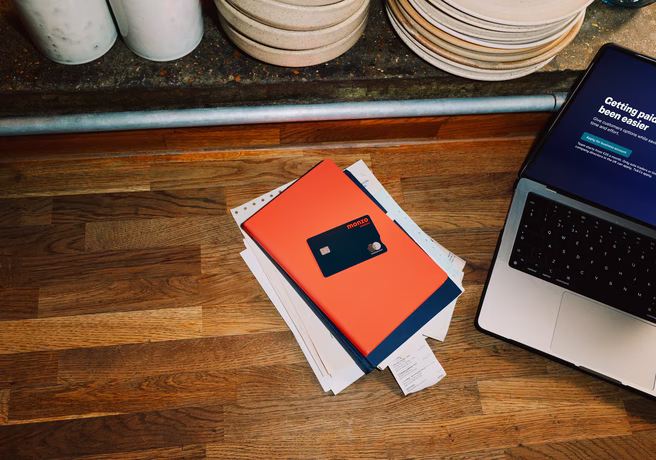 Overhead view of a restaurant workspace with a Monzo business card resting on paperwork beside a laptop, stacked plates visible on the shelf above.