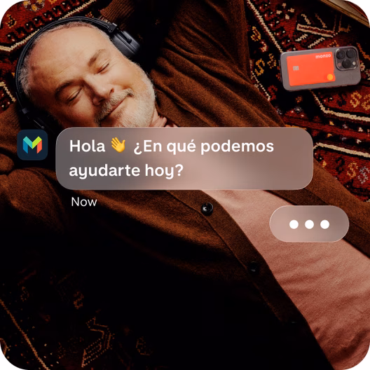Hombre relajándose y escuchando música con una tarjeta de color coral de Monzo a su lado y un mensaje del servicio de atención al cliente de Monzo en la app que pregunta: «¿Cómo podemos ayudarte hoy?».