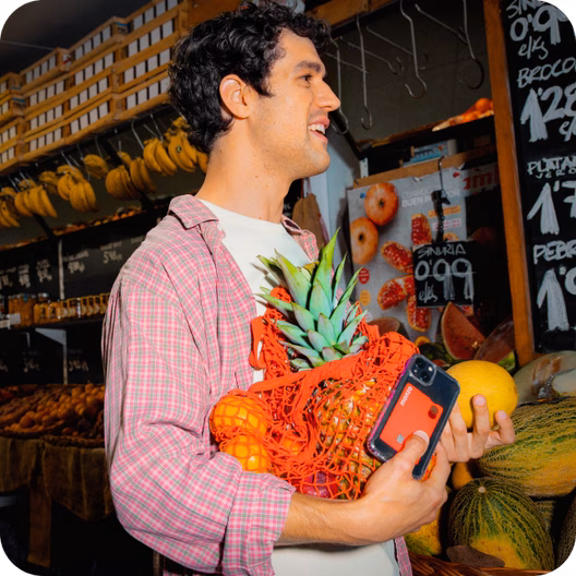 Hombre sonriendo en un mercado mientras sostiene una bolsa con la compra y una tarjeta de color coral de Monzo.