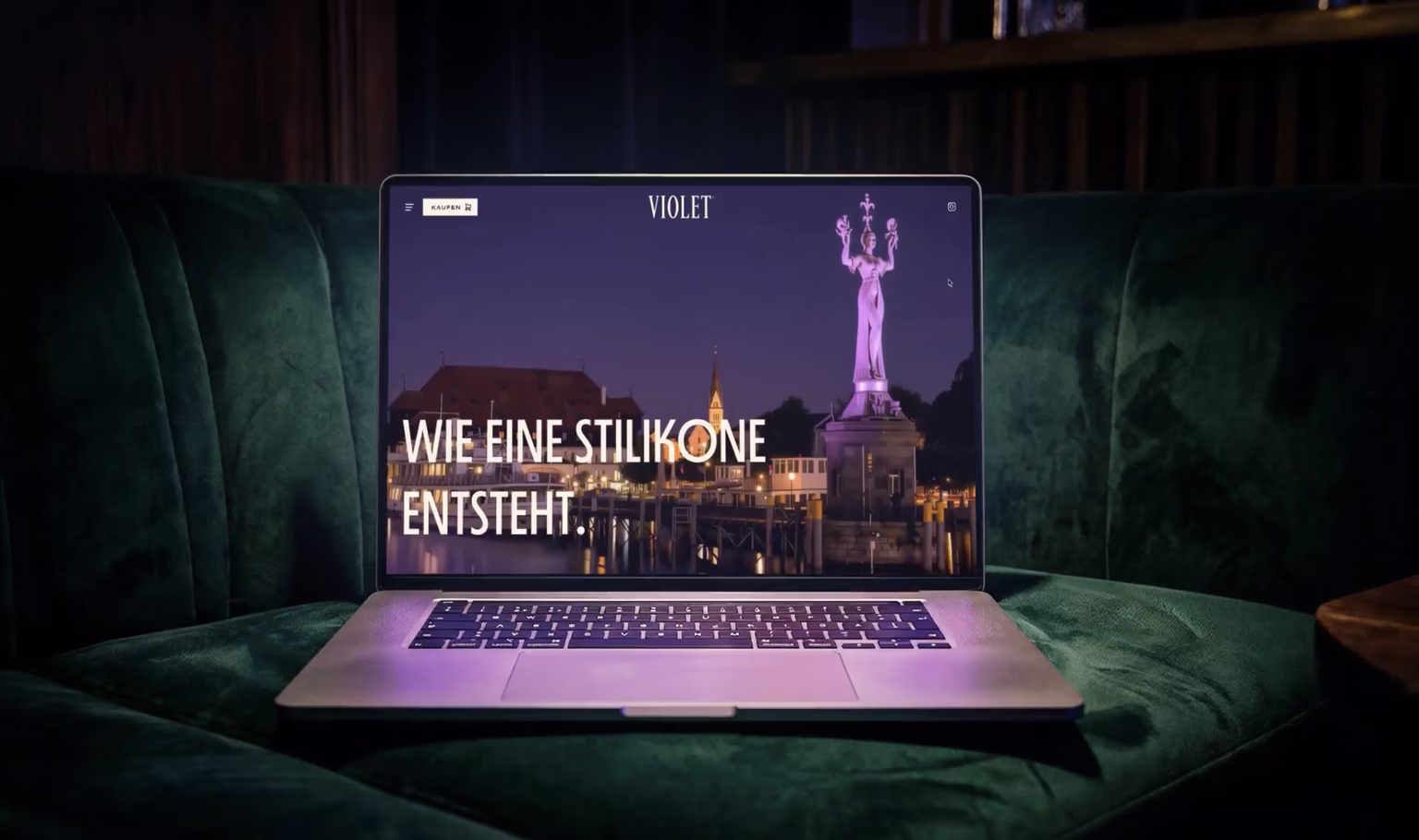 Laptop on a green velvet couch shows a purple webpage titled "VIOLET," featuring a statue monument against a cityscape.