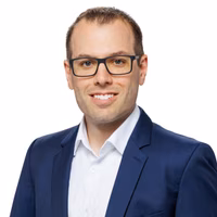 Porträt eines Mannes im Anzug mit Brille, kurzem Haar und weißem Hemd. Er lächelt freundlich vor weißem Hintergrund.