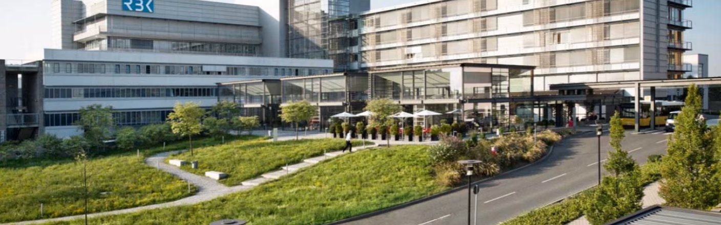 Modernes Gebäude mit Glasfassade und Rasenfläche davor; Spazierwege und Bäume, mit einer Straße am rechten Bildrand.