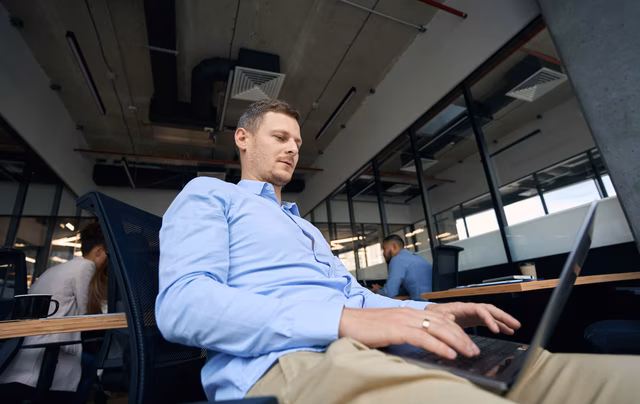 Konzentrierter Unternehmensmitarbeiter, der auf einem Stuhl sitzt und an seinem Laptop mobil mit seinen Bürokollegen arbeitet
