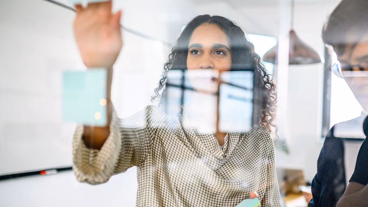 Person schreibt auf Haftnotizen an einer Glasscheibe in einem Büro und leitet einen Workshop.