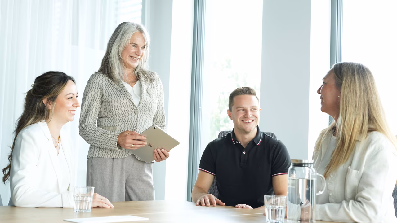 Vier Personen sitzen an einem Tisch in einem modernen Büro. Eine Frau steht und hält ein Tablet.