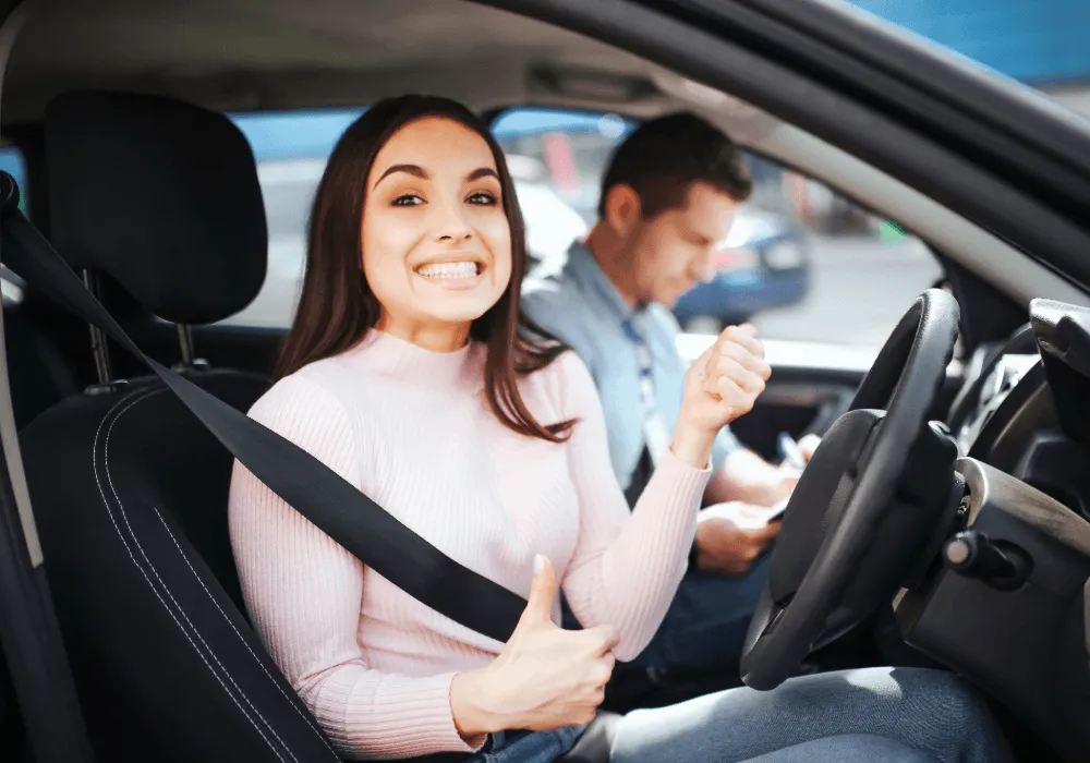 Smiling woman in driver’s seat gives a thumbs up while wearing a seatbelt, with an instructor checking notes beside her.