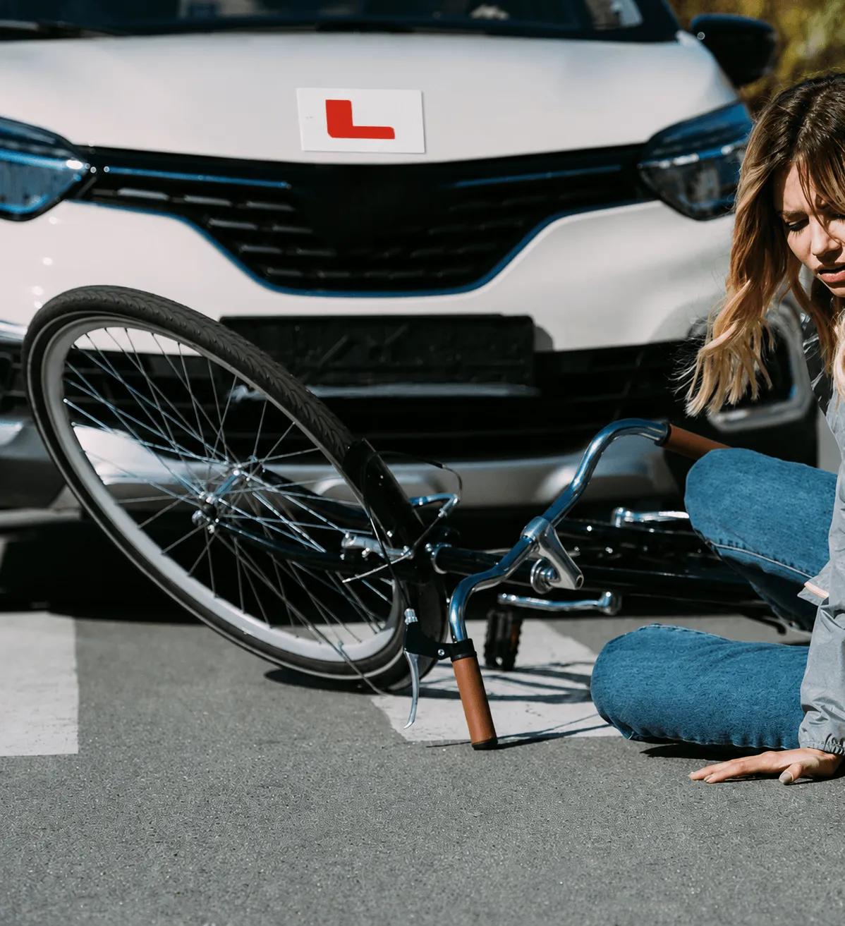 A distressed cyclist sitting on the road next to a fallen bicycle directly in front of a white driving school car displaying an L-plate.