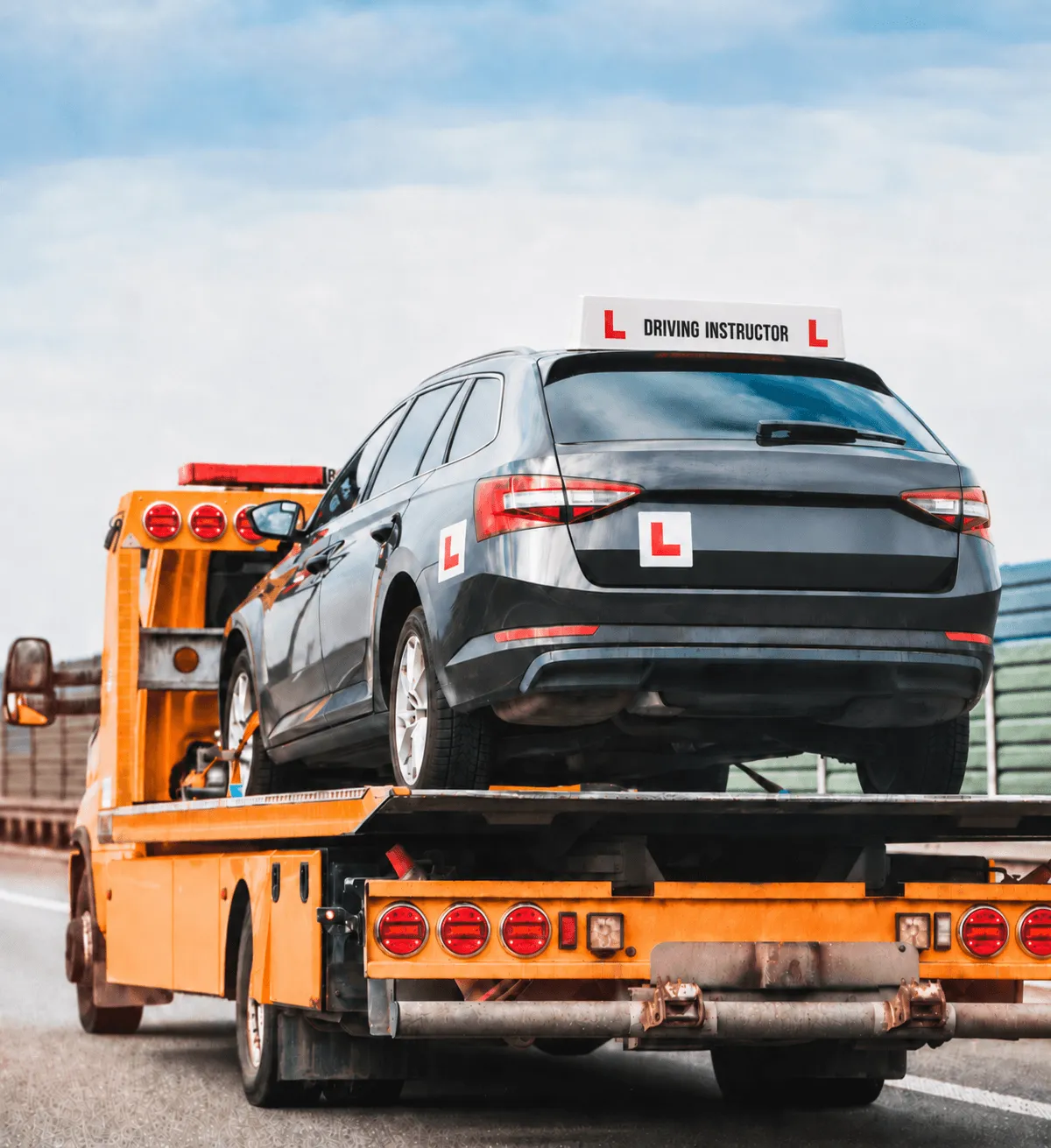 A black driving instructor car with L-plates being transported on the back of a yellow flatbed recovery truck.