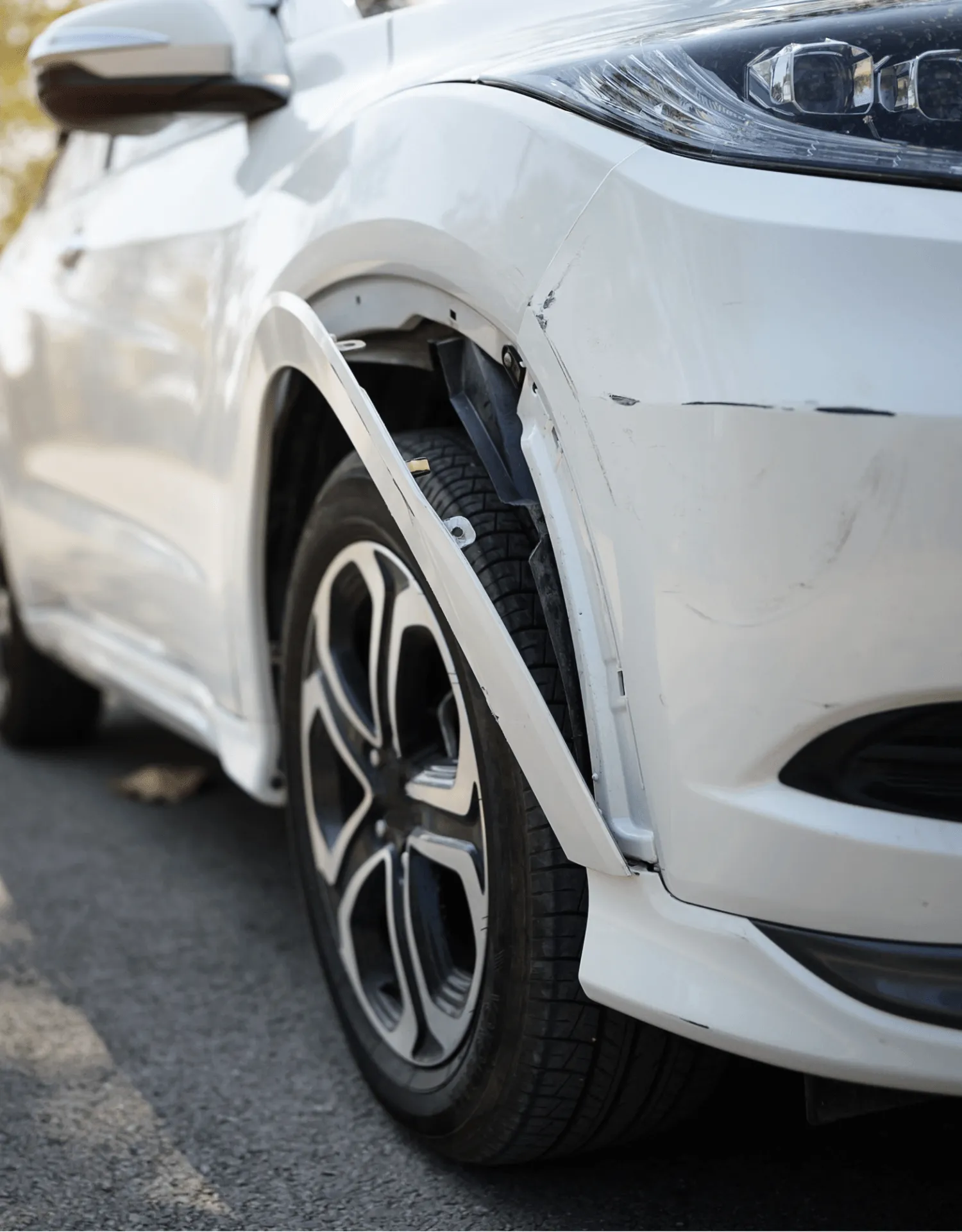 A close-up of a white car showing collision damage, with scuff marks on the front bumper and the plastic wheel arch trim broken and hanging loose over the tyre.