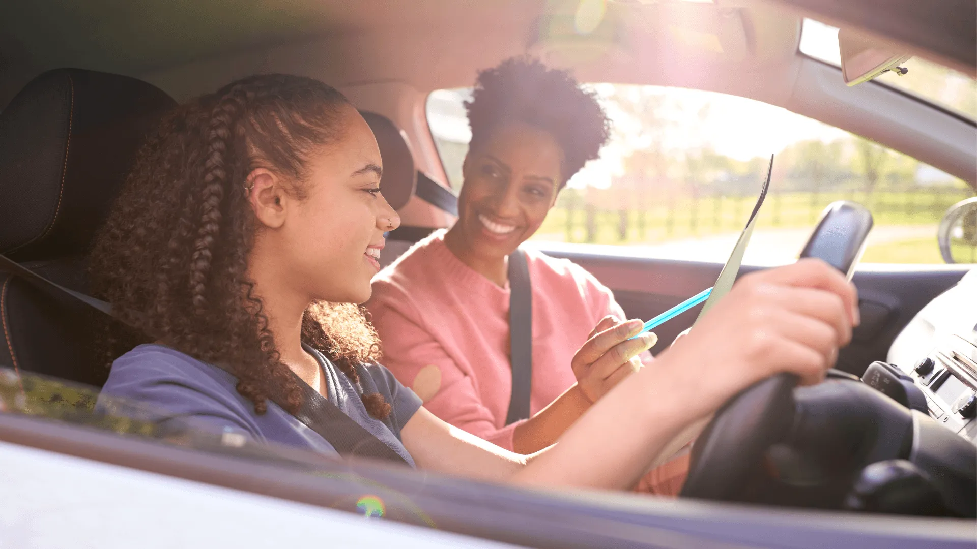 A teen girl at the steering wheel smiles as an adult woman in the passenger seat reviews a clipboard during a driving lesson.