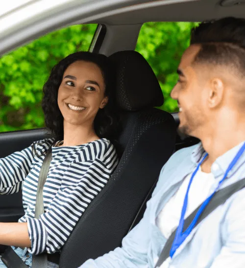 A smiling young woman driving a car, with a male instructor wearing a blue lanyard in the passenger seat.