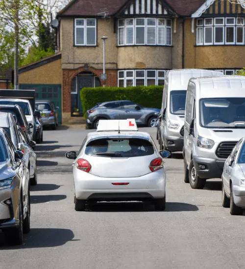 Rear view of a silver driving school car with an L-plate roof sign navigating a narrow UK residential street lined with parked vehicles.