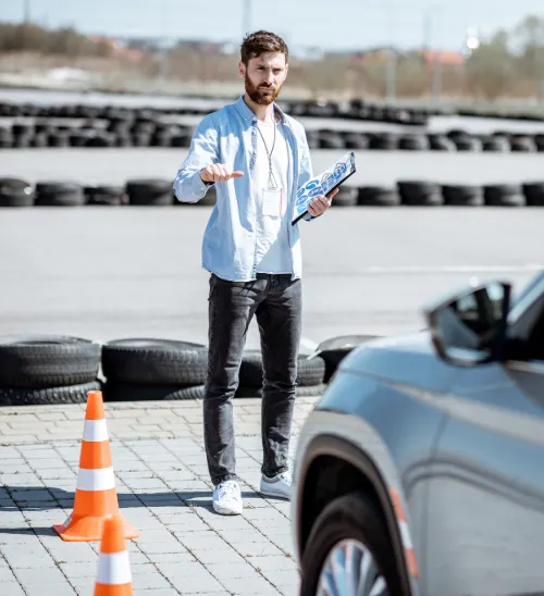 Driving instructor with clipboard signals a car to stop beside traffic cones on a test track lined with tires.
