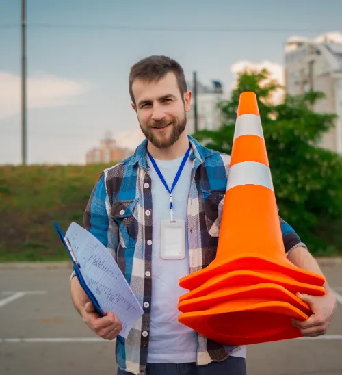 Smiling man in a plaid shirt holding stacked orange traffic cones and a clipboard in an outdoor parking lot.