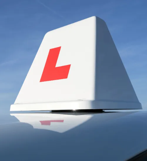A close-up of a white driving school roof sign displaying a red L-plate attached to the top of a tuition car against a blue sky.