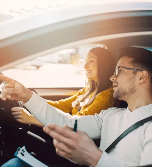 A driving instructor holding a clipboard and pointing the way for a smiling learner driver, demonstrating how courtesy vehicle cover keeps you working.