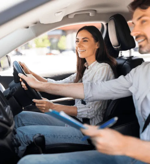 A driving instructor sitting in the passenger seat holding a clipboard while reaching over to assist a smiling female learner driver with the steering wheel.