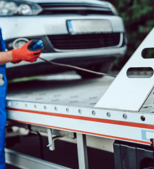 A roadside recovery worker in blue overalls and red gloves using a winch control to safely load a broken-down silver car onto a flatbed tow truck.