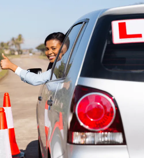 A smiling learner driver gives a thumbs-up out the window of a silver tuition car with L-plates next to traffic cones.