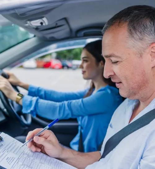 Driving instructor fills out a form while a young woman in the driver’s seat practices driving in a parked car.