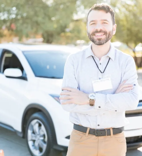 Smiling driving instructor with folded arms stands in front of a white SUV in a sunny outdoor setting.