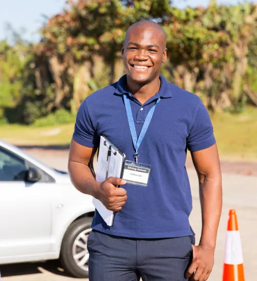 Smiling driving instructor in a navy polo holds a clipboard near a white car in an outdoor training area.