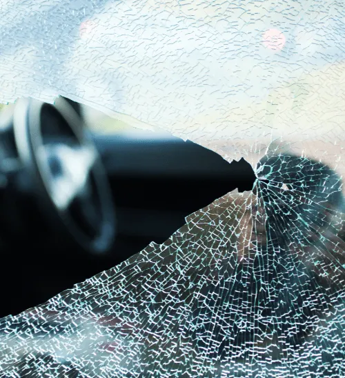 Shattered car windshield with a central impact point, showing spider web cracks, dashboard and steering wheel visible in background.