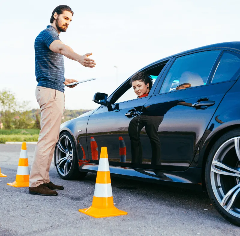 Driving instructor with clipboard gesturing to female student in black car surrounded by orange traffic cones during driving lesson.