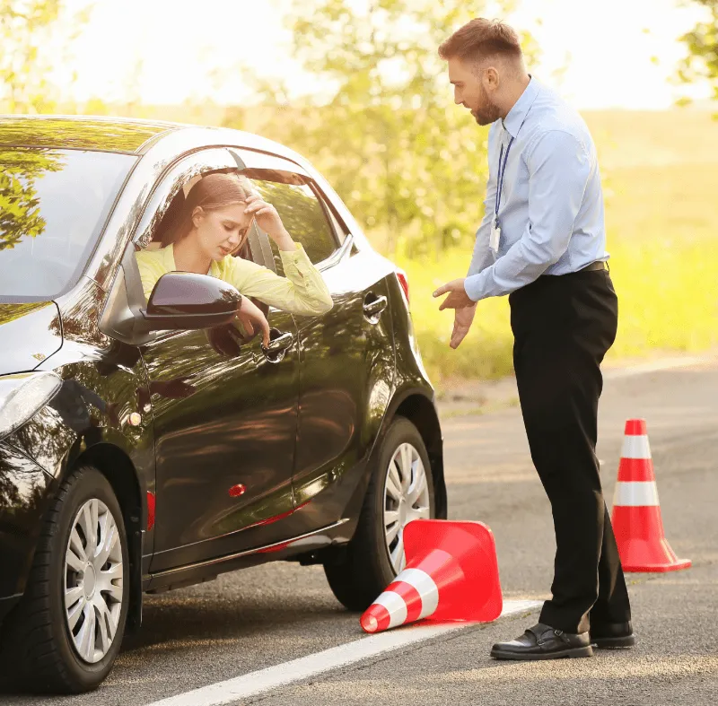 A driving instructor gestures at a knocked-over traffic cone while a stressed female student sits in a black car during a driving test.