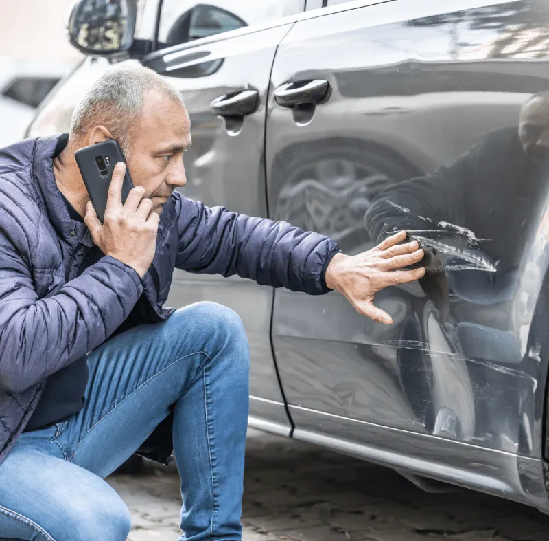 Man on phone crouches beside a car, inspecting a large scrape and dent on the vehicle’s side door.