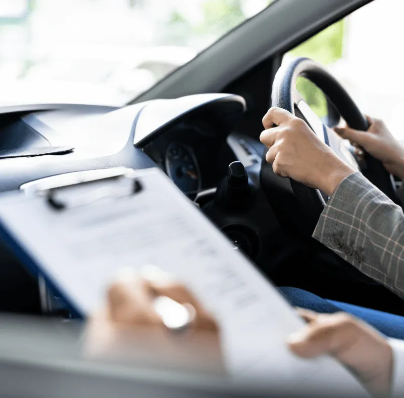 Driving instructor holding clipboard while student grips steering wheel during a driving lesson or test inside a car.