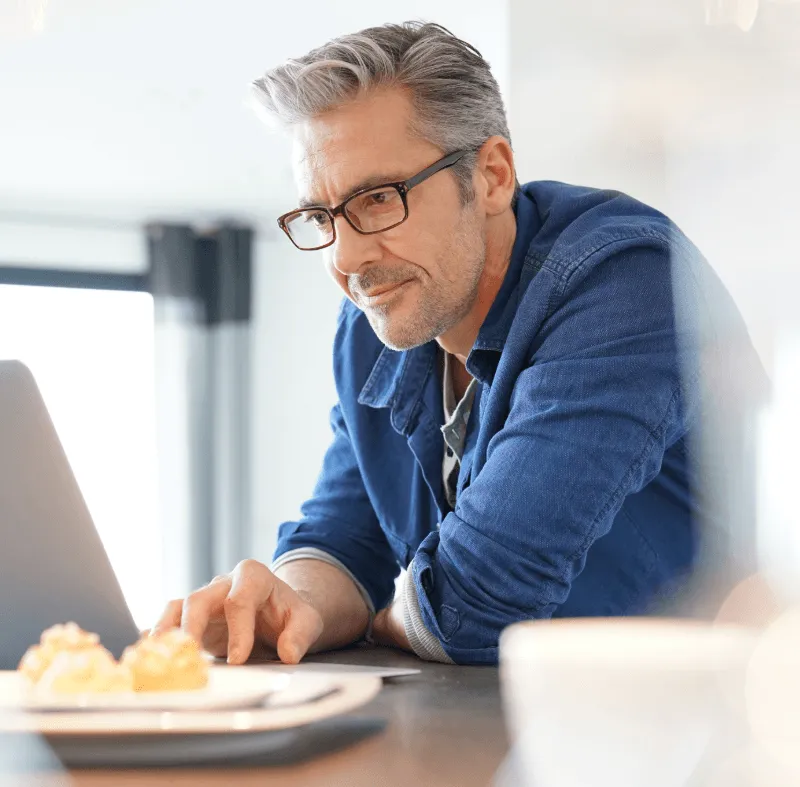 Middle‑aged man in glasses and denim shirt working on a laptop at a bright kitchen table with pastries in the foreground.