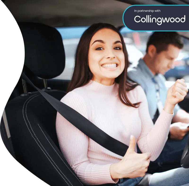 Smiling woman in the driver’s seat gives a thumbs up while wearing a seat belt, with a man in the passenger seat in the background.