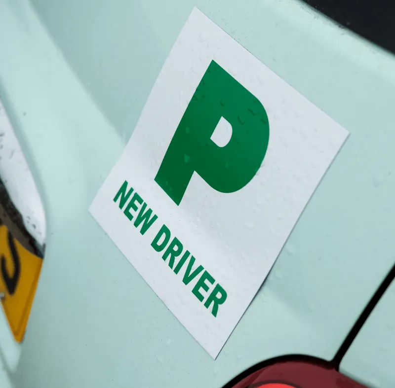 Close-up of a car with a green “P NEW DRIVER” sign on the wet rear panel.