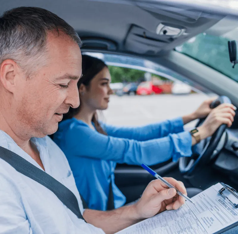 A driving instructor marks a clipboard while a young woman in blue drives during a road test.
