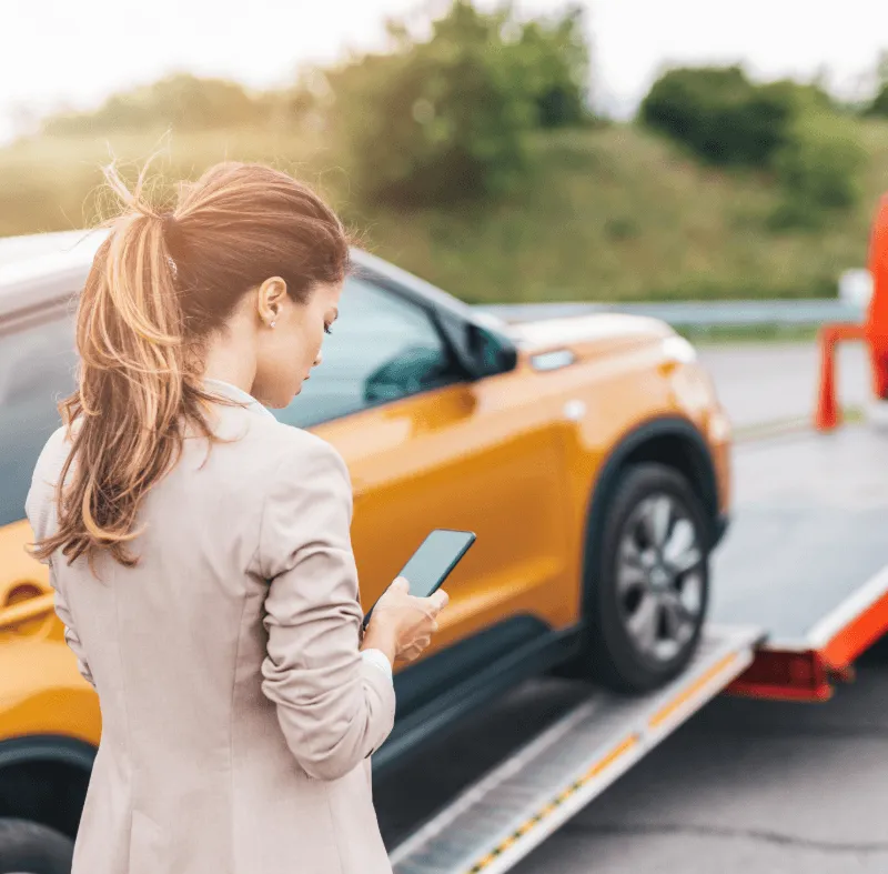 Woman using smartphone beside yellow SUV being loaded onto a tow truck on a roadside.
