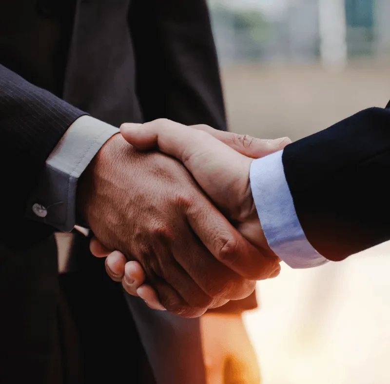Two business professionals in suits shaking hands, symbolizing agreement or partnership, with a blurred urban background.