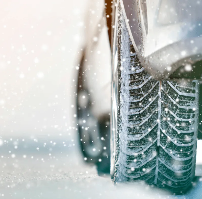 Close-up of a car tire with snow-covered tread driving on a snowy road with falling snowflakes.