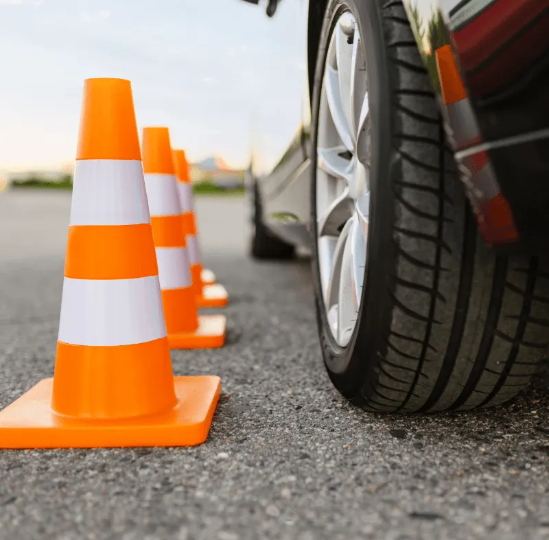 A row of orange traffic cones with white stripes alongside a car tire on an asphalt surface, suggesting a driving course or road safety setup.