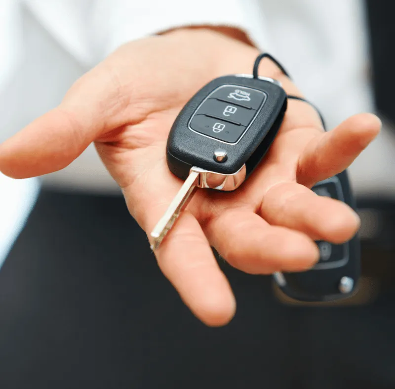 A hand holding a black car key fob with a flip-out metal key and three buttons, suggesting a car sale or rental.