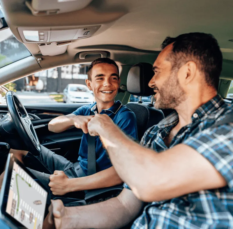 A smiling teen driver fist-bumping an adult instructor inside a car, with a GPS tablet visible, during a driving lesson.