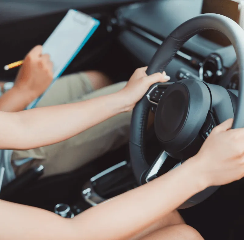 Learner driver holding the steering wheel while a driving instructor writes notes on a clipboard, representing specialist insurance for dual control cars.