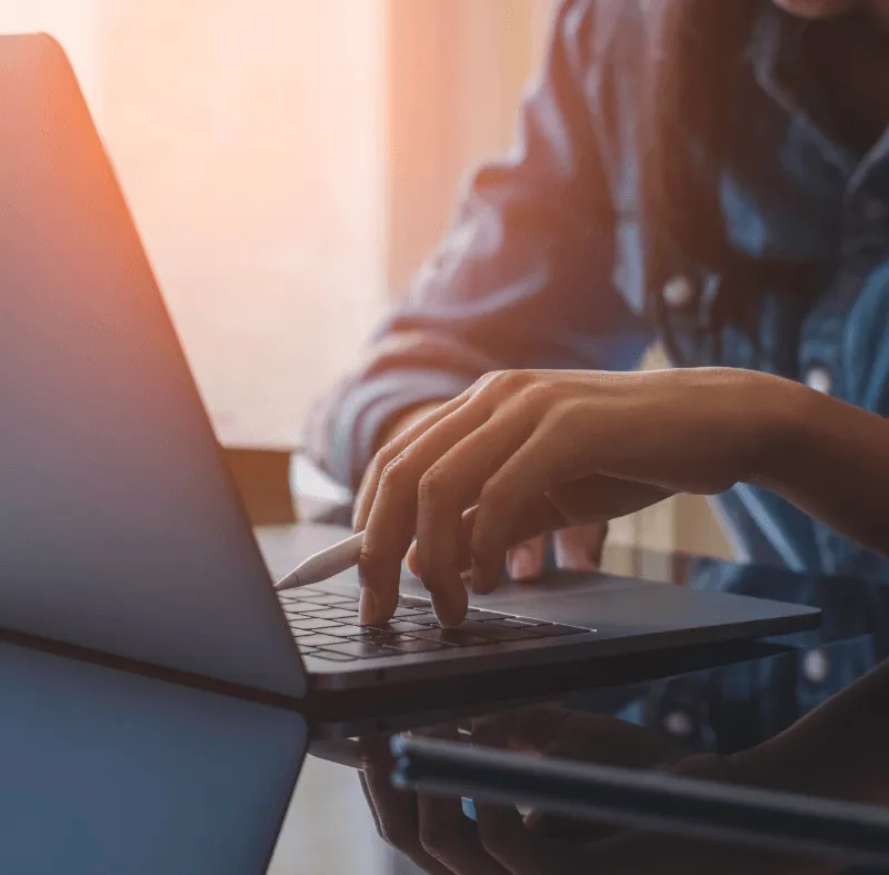 Close-up of a person’s hand using a stylus and typing on a laptop at a reflective desk in warm light.