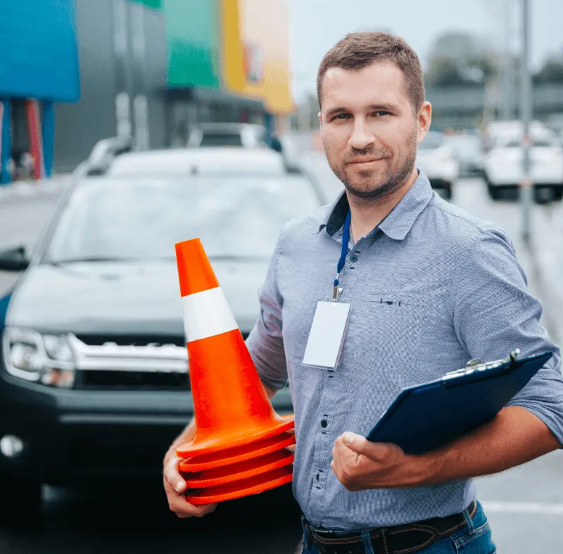 Driving instructor holding traffic cones and a clipboard beside a learner car, representing named driver and any driver insurance policies.