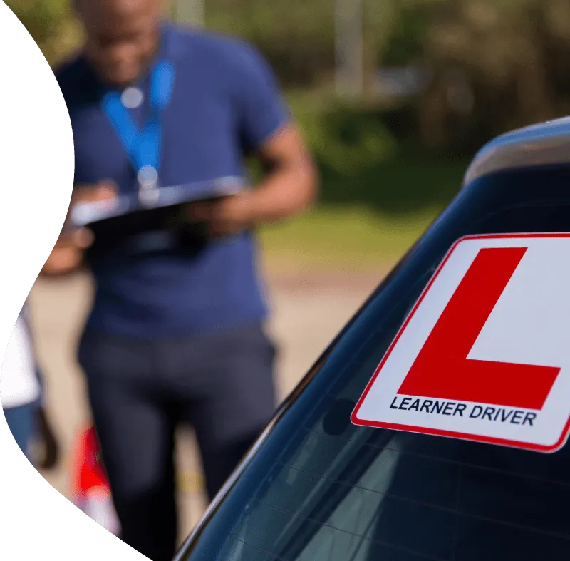 Learner driver car with a red L plate on the window, instructor holding a clipboard blurred in the background.
