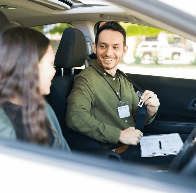 Driving instructor fastening seat belt and smiling at a student in the passenger seat during a lesson.